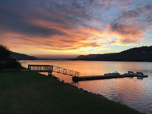 Puesta de sol sobre un lago con muelle y pequeñas embarcaciones, rodeado de montañas.