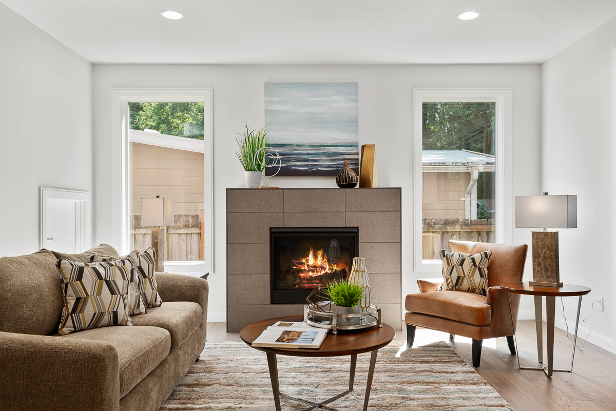 Living room with fireplace, plush sofa, armchair, round tables, plants, and large windows overlooking outdoors.